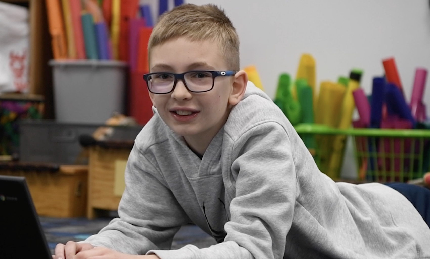 Boy with glasses smiles at the camera while laying on his stomach while and typing on his computer.