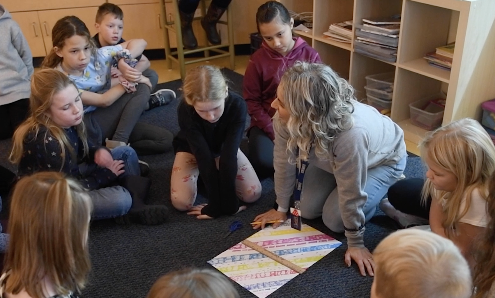 Kids all sitting on the floor, surrounding their art teaching, while she draws on a paper.